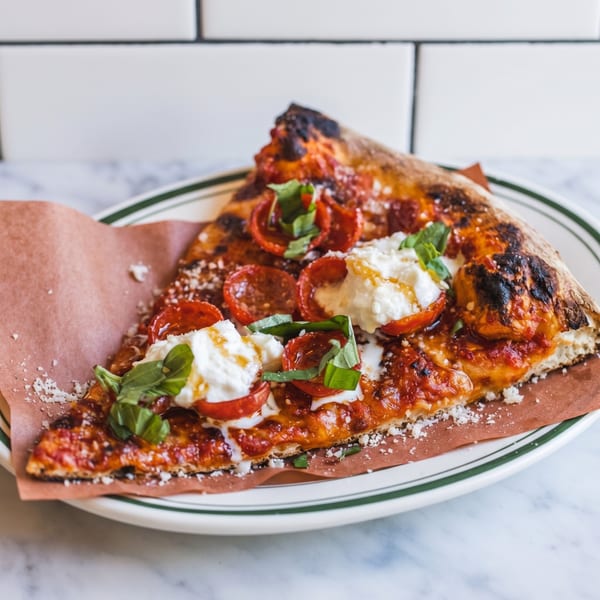 A slice of pizza with pepperoni, burrata, basil, and a charred crust on a white plate.