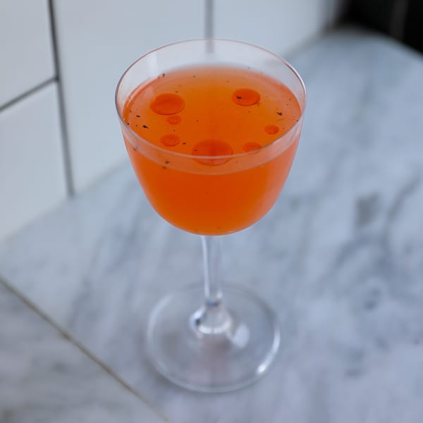 Vibrant orange cocktail in a coupe glass, garnished with small red oil drops and dark specks.