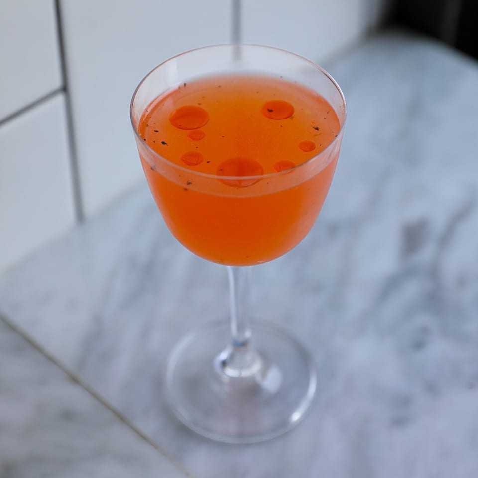 Vibrant orange cocktail in a coupe glass, garnished with small red oil drops and dark specks.