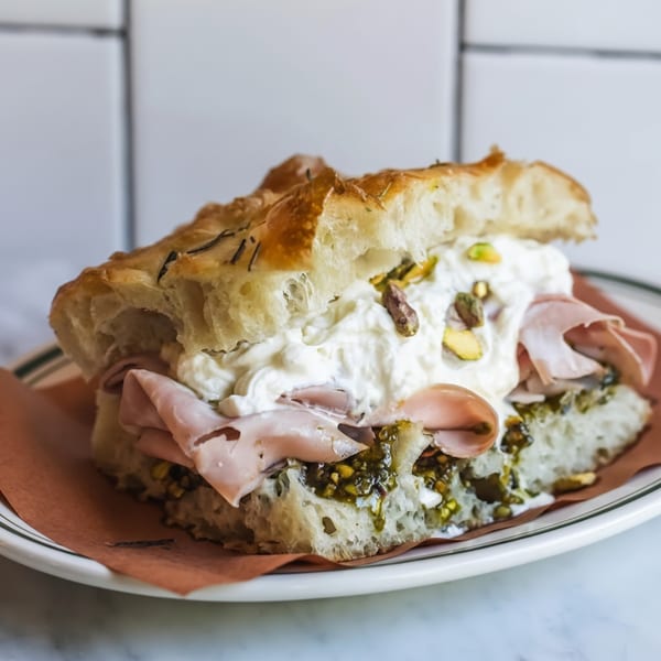 Focaccia sandwich with mortadella, burrata, pesto, and pistachios on a white plate.