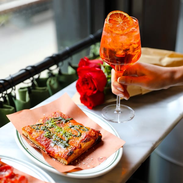 A hand holding an orange cocktail next to a slice of pizza and a red rose.