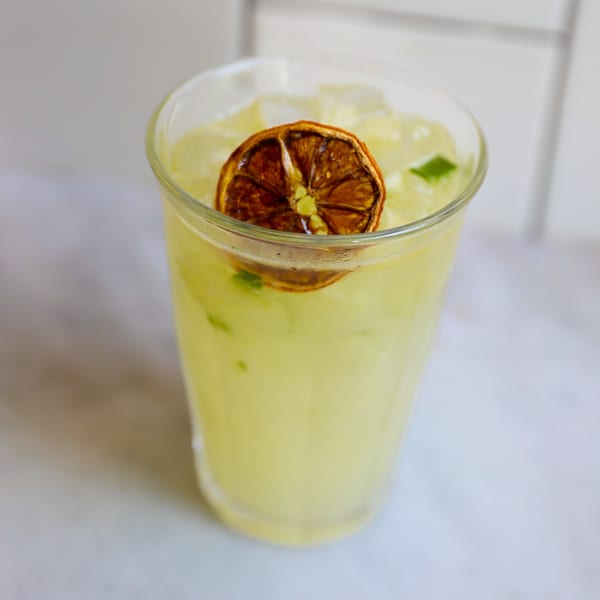 A glass of light yellow lemonade with ice, garnished with a dried orange slice and mint.
