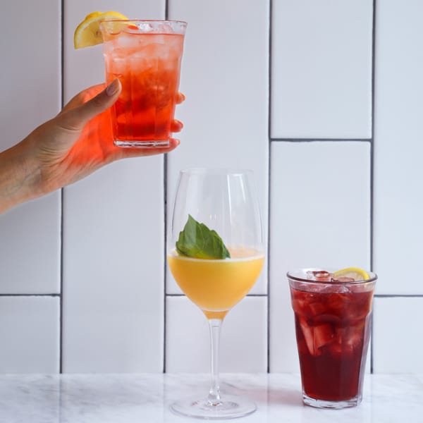 A hand holds a red drink with lemon, next to a yellow drink and another red drink.