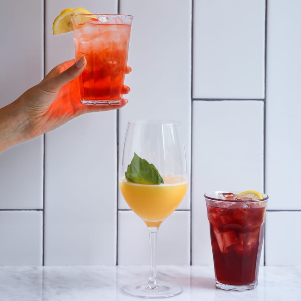 A hand holds a red drink with lemon, next to a yellow drink and another red drink.