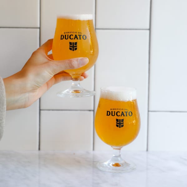 A hand holds a glass of Birrificio del Ducato beer, with another glass on a marble counter.