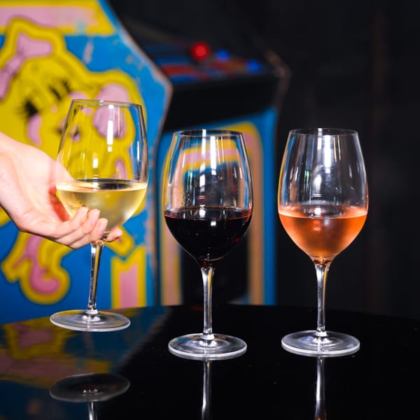 A hand holds a glass of white wine next to red and rosé wines on a table with an arcade game in the background.