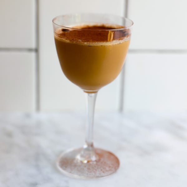 A cocktail in a stemmed glass, topped with foam and cinnamon, on a marble surface.